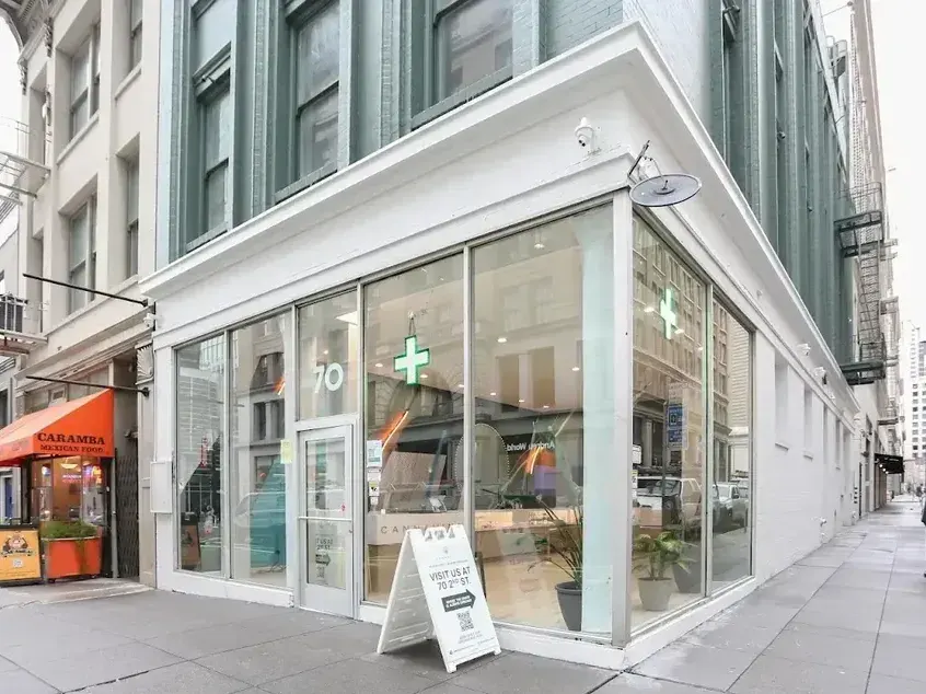 Corner storefront of a cannabis dispensary with glass windows and green cross sign